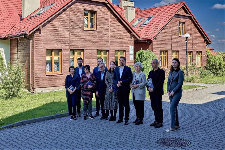 Grupa osób pozuje na stojąco do wspólnego zdjęcia przed zabudowaniami