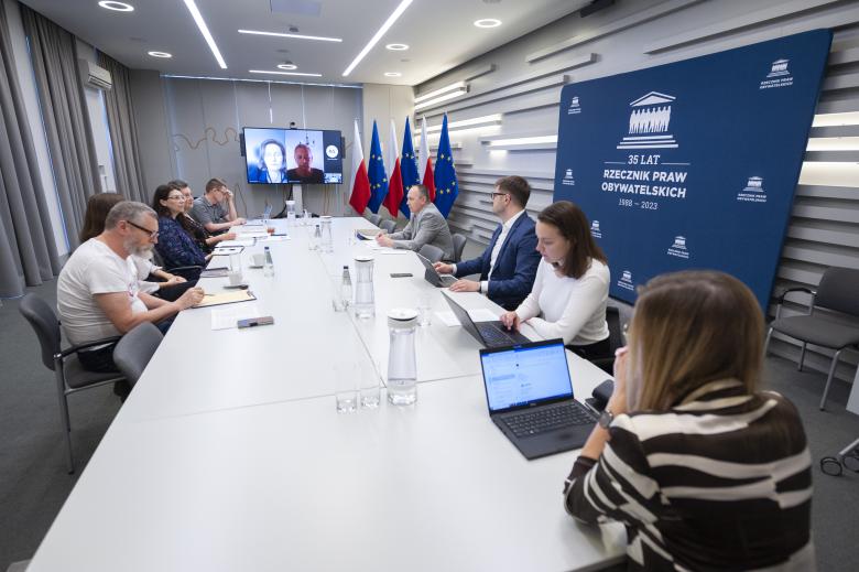 ZRPO Valeri Vachev i członkowie inicjatywy "Nasz Rzecznik" rozmawiają przy konferencyjnym stole. W szczycie stołu stoi monitor wyświetlający twarze uczestników spotkania łączących się za pośrednictwem Internetu. W tle flagi Polski i Unii Europejskiej oraz logo Rzecznika Praw Obywatelskich