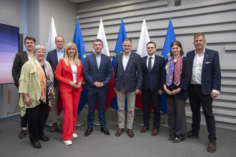 Dziewięć osób pozuje na stojąco do zdjęcia na tle flag Polski i Unii Europejskiej