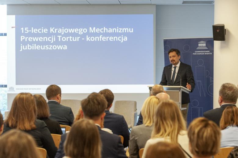 RPO Marcin Wiącek przemawia na konferencji, w tle na ekranie napis "15-lecie Krajowego Mechanizmu Prewencji Tortur - konferencja jubileuszowa"
