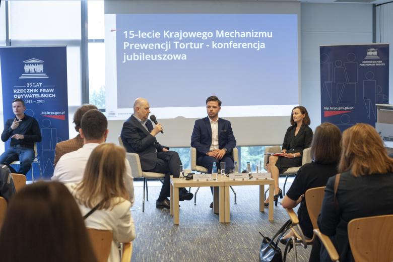 Trzy osoby siedzą na krzesłach przy stole w trakcie panelu dyskusyjnego na konferencji