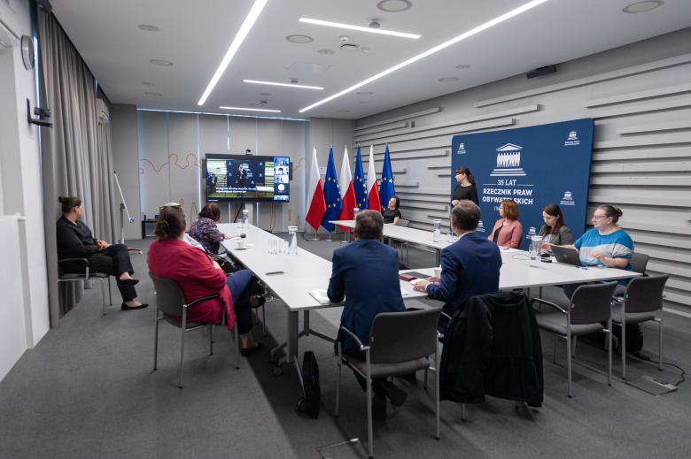 Członkowie komisji siedzą przy stole w kształcie podkowy w sali konferencyjnej. W szczycie stołu stoi ekran wyświetlający uczestników łączących się za pośrednictwem Internetu. Obok stołu stoi tłumaczka polskiego języka migowego.