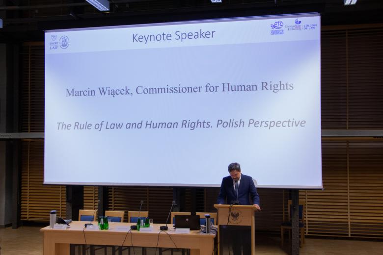 RPO Marcin Wiącek przemawia z mównicy w auli uniwersyteckiej. Za jego plecami wyświetlany jest tytuł jego wystąpienia: "The Rule of Law nad Human Rights. Polish Perspective".