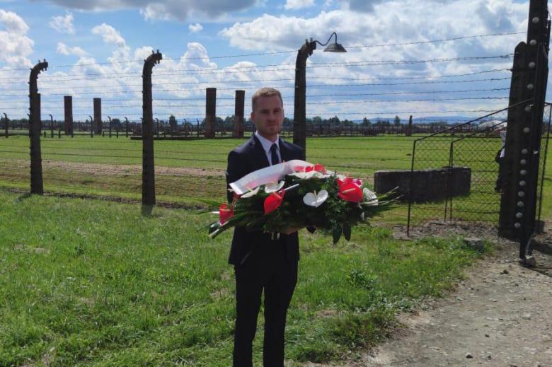 Mężczyzna w garniturze trzyma biało-czerwony wieniec na tle ogrodzenia obozowego