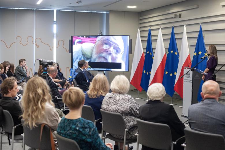 Kilkadziesiąt osób siedzących na sali konferencyjnej, którzy oglądają i słuchają nagrania wideo na dużym telewizorze