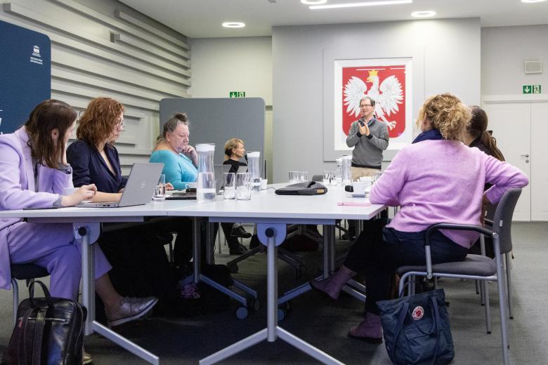 Członkowie Komisji obradują przy stole konferencyjnym. U szczytu stołu stoi mężczyzna wypowiadający się w polskim języku migowym