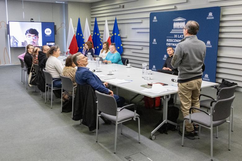 Członkowie Komisji obradują przy stole konferencyjnym. Przy jednym ze szczytów stołu stoi mężczyzna wypowiadający się w polskim języku migowym. W drugim szczycie stołu stoi ekran, na którym wyświetlany jest członek Komisji uczestniczący w obradach za pośrednictwem Internetu.