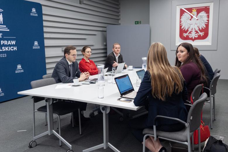 Siedem osób siedzi po obu stronach stołu i rozmawia. W tle godło Polski