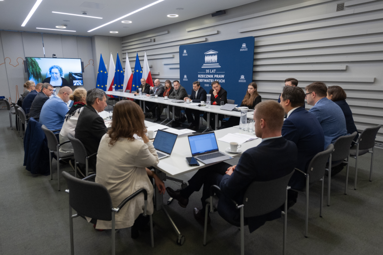 Kilkanaście osób siedzi przy stołach ustawionych w kształt podkowy na sali konferencyjnej