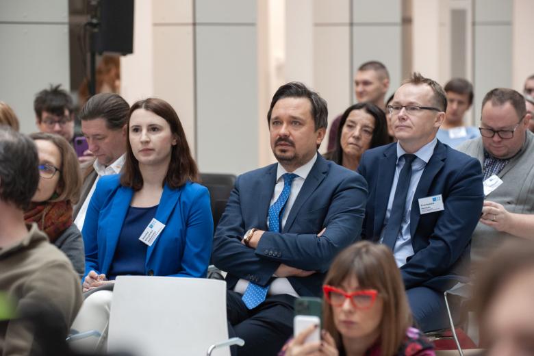 Dwie siedzące osoby w centralnej części pośród innych osób zgromadzonych na sali konferencyjnej