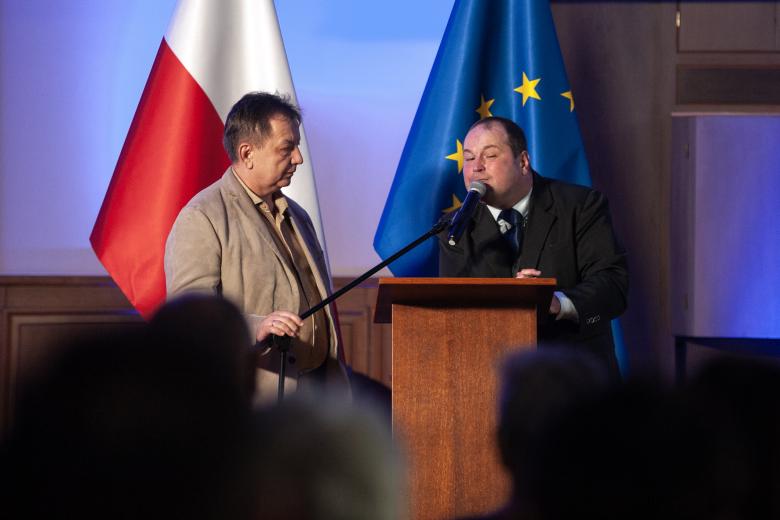 Michał Piechel wypowiada się w stronę publiczności z podium. Obok niego stoi asystująca go druga osoba. 