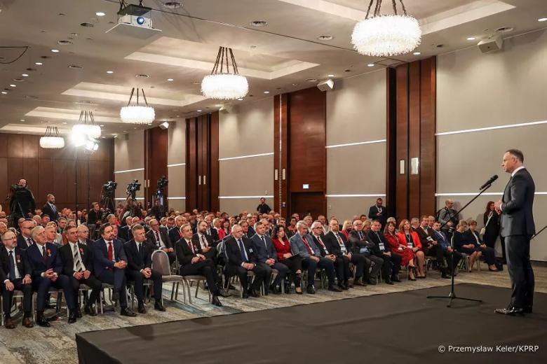 Osoba przemawia do mikrofonu na scenie, a kilkadziesiąt osób siedzących w sali konferencyjnej słucha wystąpienia