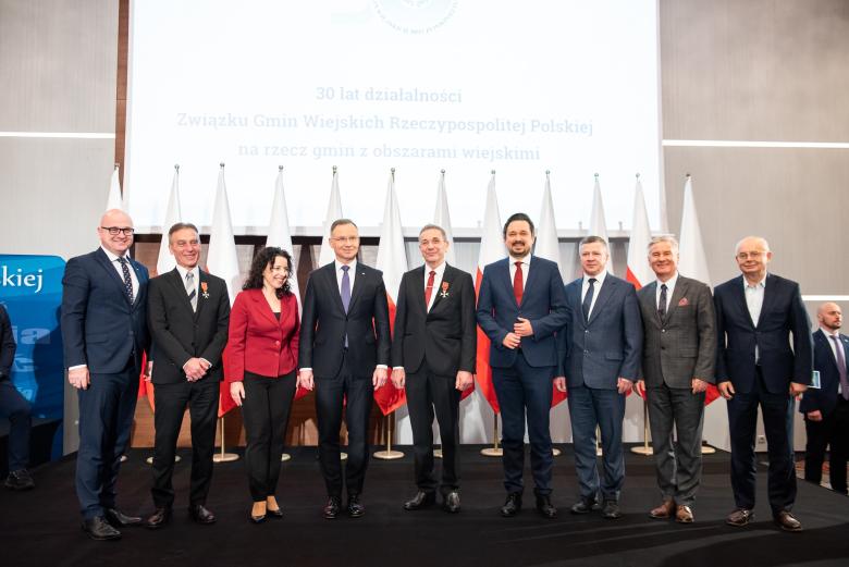 Grupa osób pozuje na stojąco do wspólnego zdjęcia na tle flag Polski
