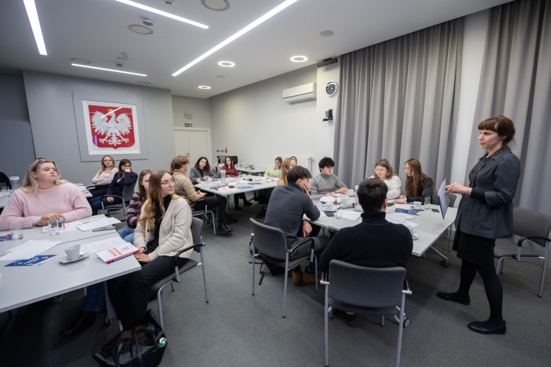 Kilkadziesiąt osób siedzących w grupach przy stołach w sali konferencyjnej słucha prowadzącego spotkanie