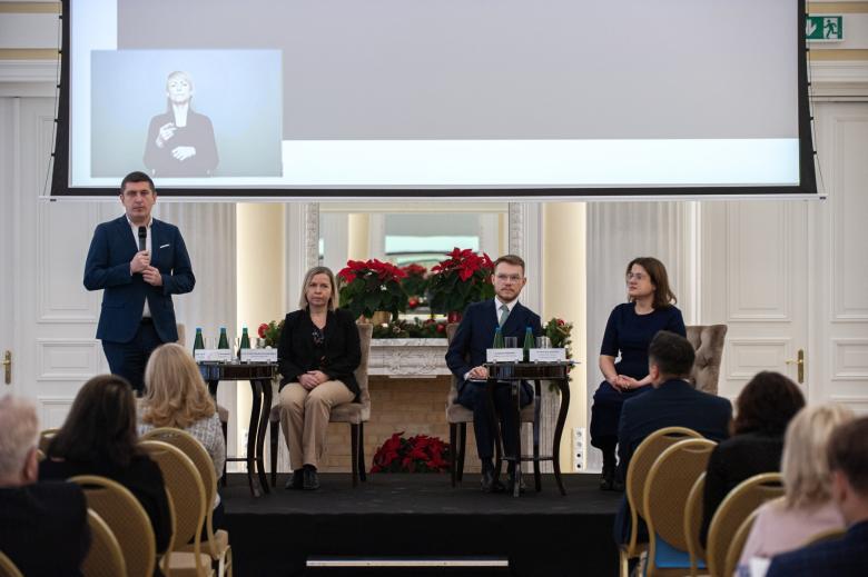 Trzy osoby siedzą na krzesłach na scenie w sali konferencyjnej, a jedna mówi na stojąco do mikrofonu