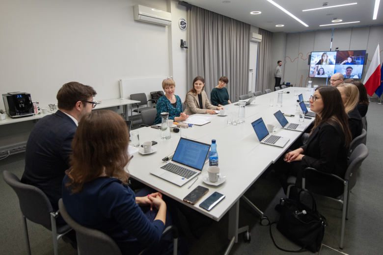 Kilkanaście osób siedzi przy stole konferencyjnym i rozmawia, w tle ekran telewizora z widocznymi w aplikacji do komunikowania się na odległość uczestnikami spotkania 