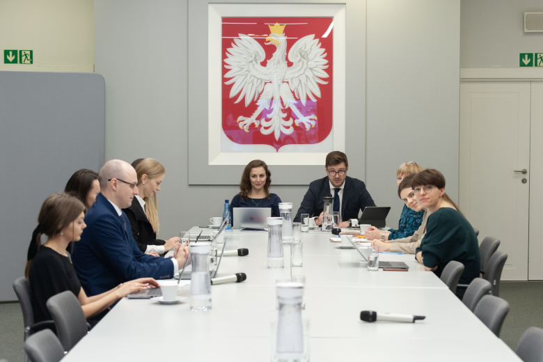 Kilkanaście osób siedzi przy stole konferencyjnym i rozmawia, w tle godło Polski z białym orłem na czerwonym tle