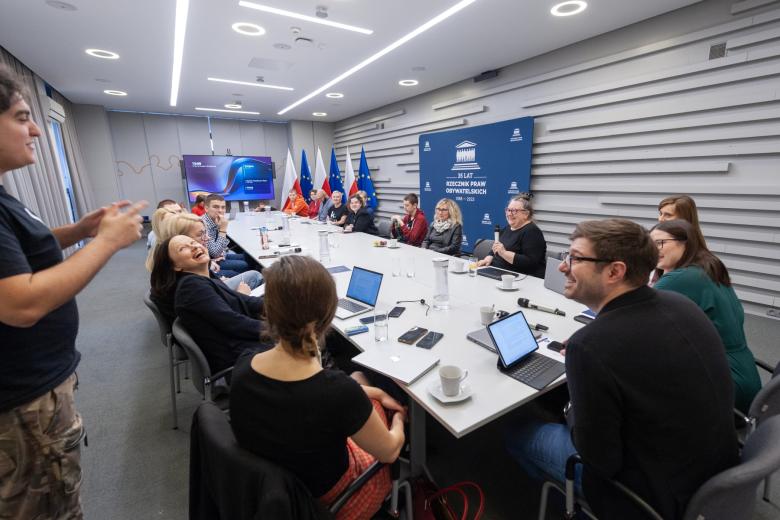 Kilkanaście uśmiechniętych osób siedzących przy dużym prostokątnym stole w sali konferencyjnych