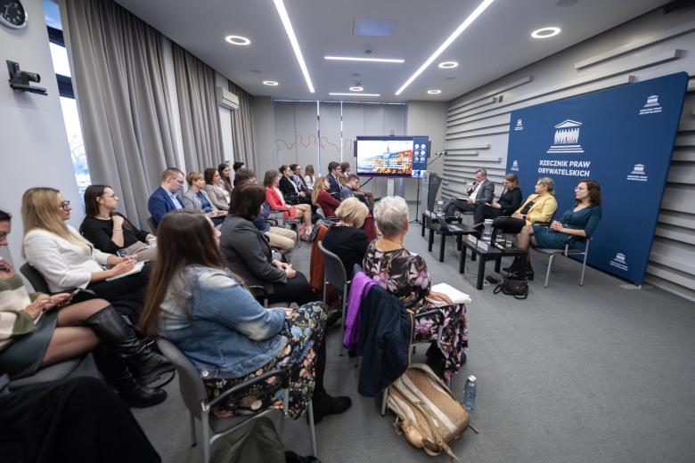 Kilkadziesiąt osób siedzi na sali konferencyjnej i słucha rozmowy czterech osób