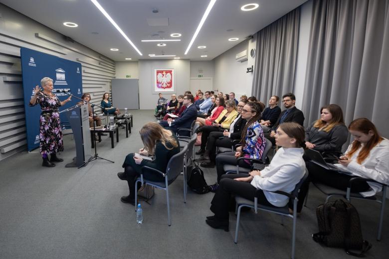 Kilkadziesiąt osób na sali konferencyjnej słucha wystąpienia osoby mówiącej za mównicą