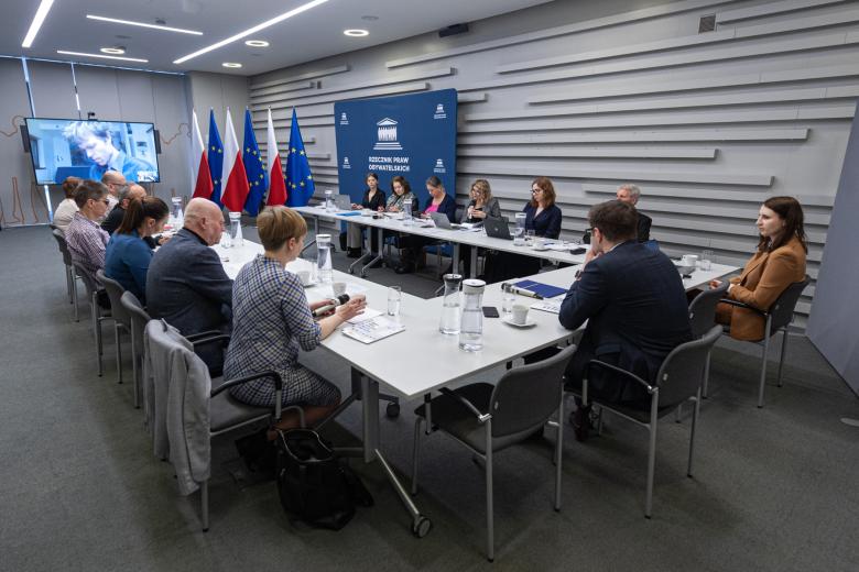 Kilkanaście osób siedzi przy stole konferencyjnym i rozmawia, w tle flagi Polski i UE