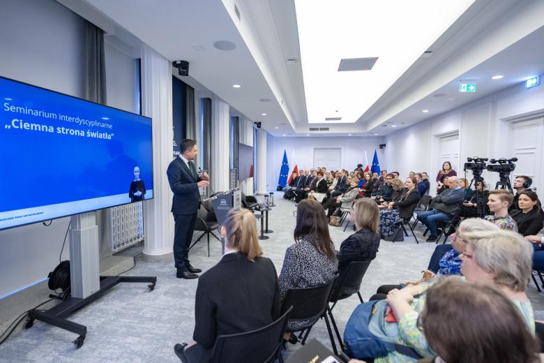 Kilkadziesiąt osób siedzących na sali konferencyjnej słucha wystąpienia osoby mówiącej za mównicą