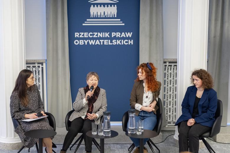 Cztery osoby siedzą na krzesłach w sali konferencyjnej, jedna mówi do mikrofonu, pozostałe słuchają, w tle granatowa plansza z białym napisem "Rzecznik Praw Obywatelskich"