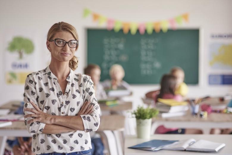 nauczycielka ze skrzyżowanymi rękami w tle klasa szkolna z dziećmi 