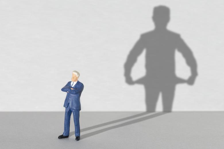 Ilustracja - figurka mężczyzny w garniturze stojącego z założonymi rękoma, która rzuca długi cień. Cień ma inny kształt, niż figurka - przedstawia osobę wywracającą na lewą stronę puste kieszenie.