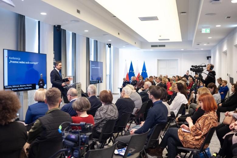 Kilkadziesiąt osób siedzących na sali konferencyjnej słucha wystąpienia osoby mówiącej za mównicą