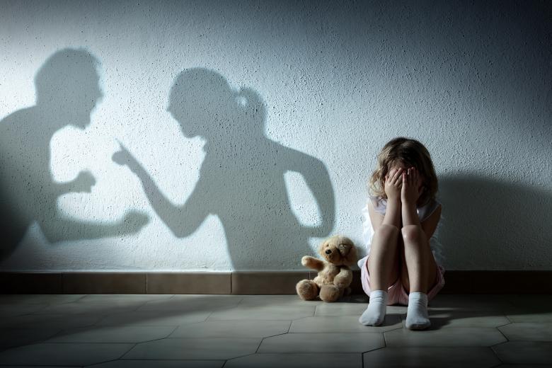 Dziewczyna siedzi przy ścianie z głową schowaną w dłoniach, obok miś pluszowy, na ścianie cień dwóch osób które rozmawiają mocno gestykulując