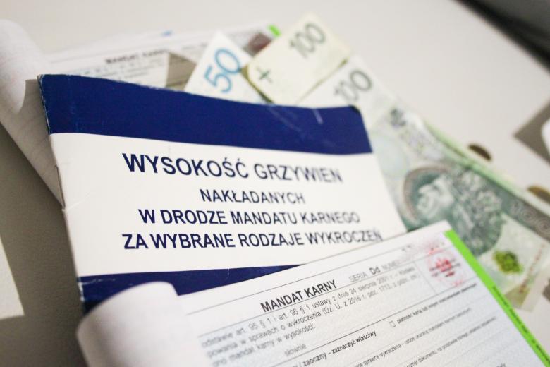 Plik druczków urzędowych do wystawiania mandatów karnych i książeczka o tytule "Wysokość grzywien nakładanych w drodze mandatu karnego za wybrane rodzaje wykroczeń" leżące na banknotach