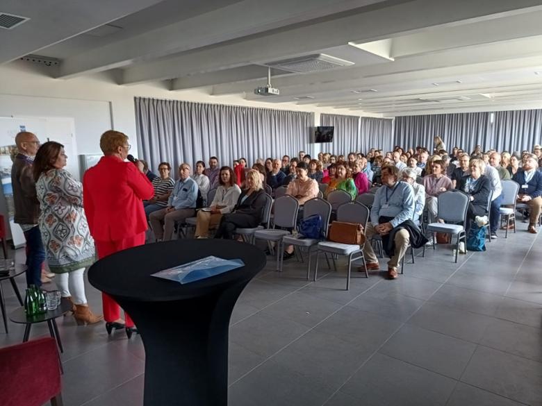 Kilkadziesiąt osób siedzących na sali konferencyjnej słucha wystąpienia osób mówiących na scenie