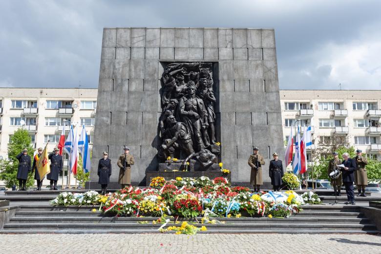 Duży pomnik przed którym leży kilkadziesiąt bukietów i wieńców kwiatów