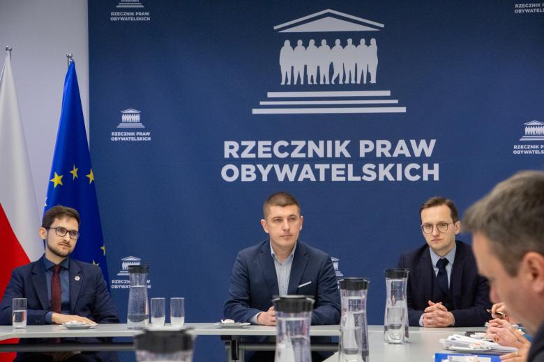 Trzy osoby siedzą za stołem, w tle granatowa ścianka z białym napisem "Rzecznik Praw Obywatelskich"