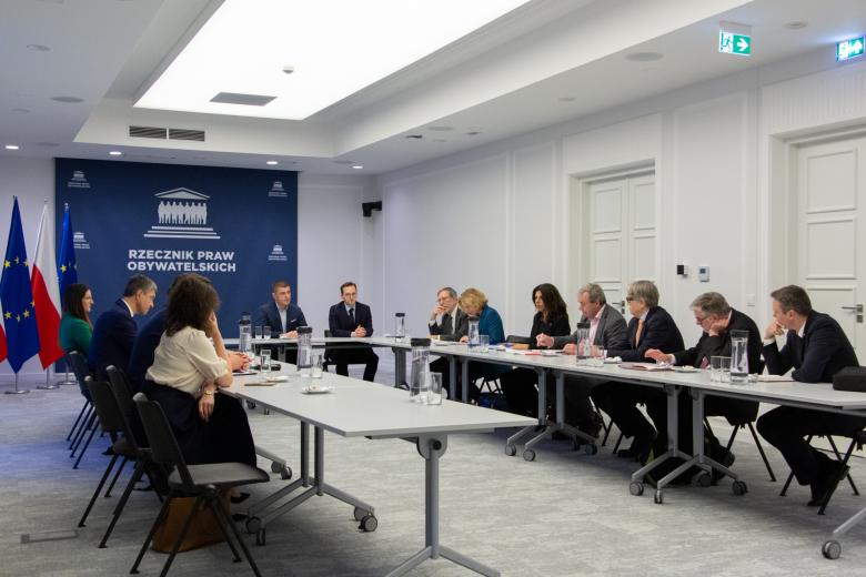 Dwanaście osób siedzi za stołami ustawionymi w podkowę na dużej sali konferencyjnej, w tle granatowa ścianka z białym napisem "Rzecznik Praw Obywatelskich"