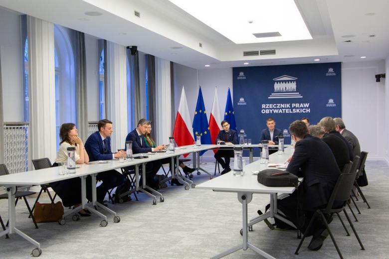 Dwanaście osób siedzi za stołami ustawionymi w podkowę na dużej sali konferencyjnej, w tle granatowa ścianka z białym napisem "Rzecznik Praw Obywatelskich"
