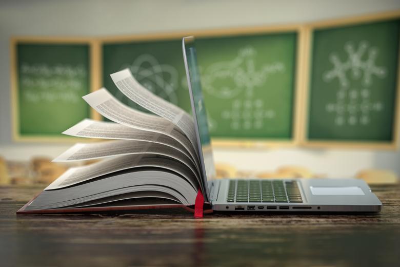 Otwarty podręcznik i laptop stoją na ławce szkolnej. W tle rozmyty widok tablicy z wzorami chemicznymi