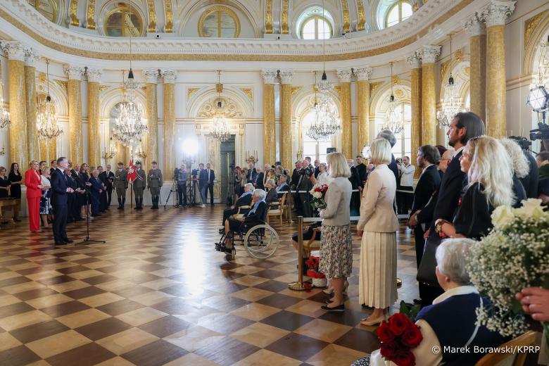 Widok ogólny dużej, bogato zdobionej sali. W centrum sali siedzą dwie osoby z założonymi orderami. Naprzeciwko stoi prezydent RP Andrzej Duda i wypowiada się do mikrofonu. Wokół stoją inni uczestnicy uroczystości