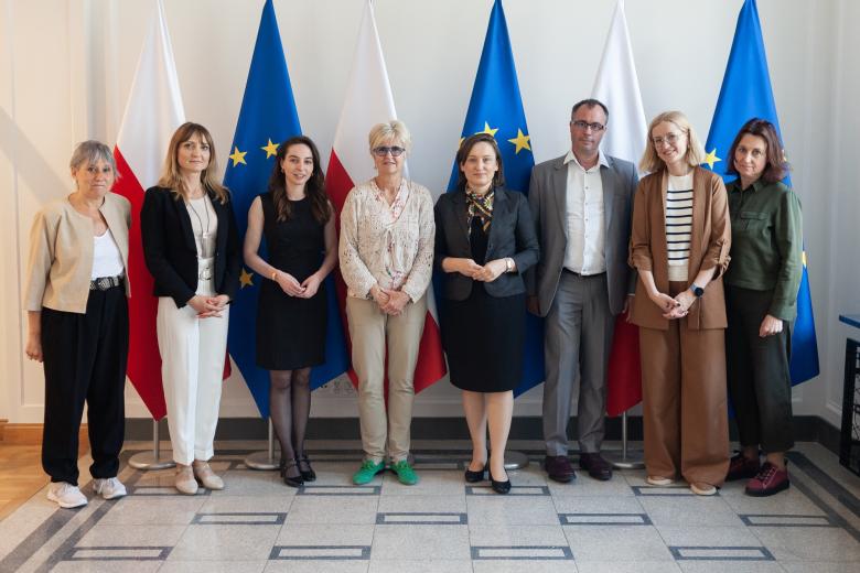 Osiem osób pozuje na stojąco do wspólnego zdjęcia na tle flag Polski i UE