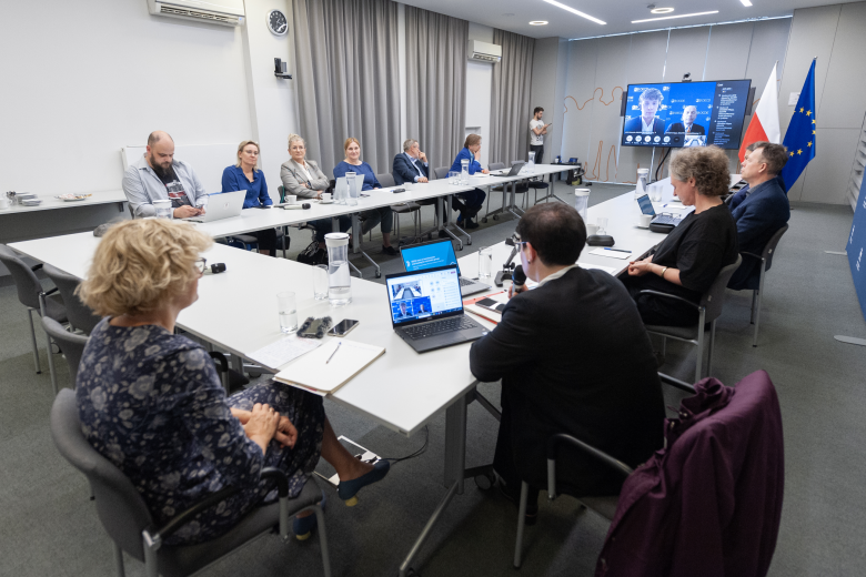 W sali konferencyjnej członkowie komisji siedzą przy ustawionym w kształt podkowy konferencyjnym stole. Na ekranie znajdującym się w szczycie stołu konferencyjnego wyświetleni są również członkowie Komisji biorący udział w spotkaniu za pośrednictwem Internetu. W tle flagi Polski i Unii Europejskiej