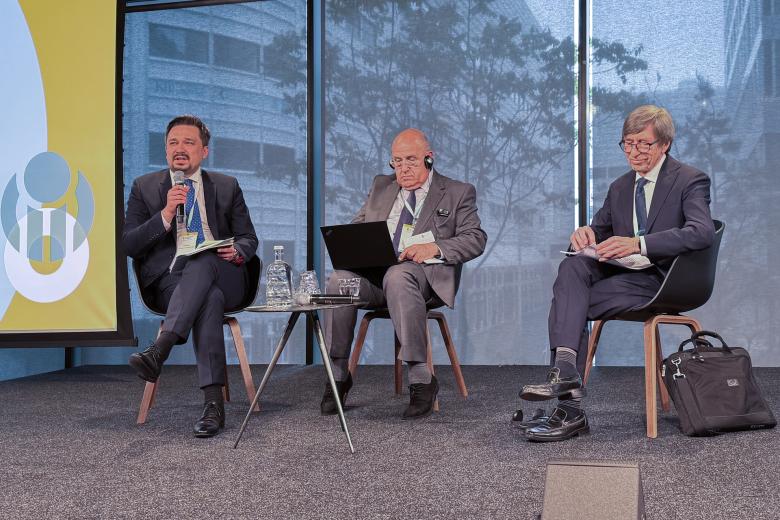 Marcin Wiącek wypowiada się do mikrofonu siedząc na ustawionym na podeście krześle. Obok niego siedzi dwóch innych mężczyzn uczestniczących w panelu