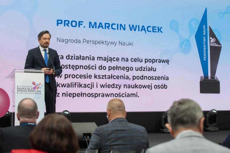 Marcin Wiącek przemawia za mównicą na scenie, w tle na dużym wyświetlaczu napis "prof. Marcin Wiącek, Nagroda Perspektywy Nauki"