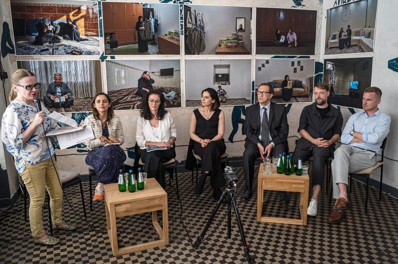 Siedem osób siedzi na krzesłach, przy małych stolikach w pomieszczeniu, w którym na ścianach wiszą fotografie