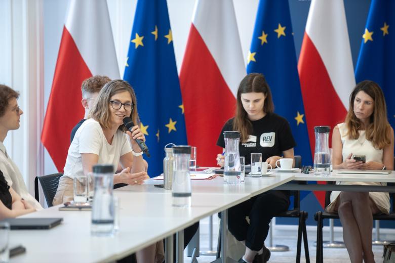 Kobieta siedząca za stołem mówi do trzymanego w dłoni mikrofonu, obok przy stole siedzą inni uczestnicy konferencji