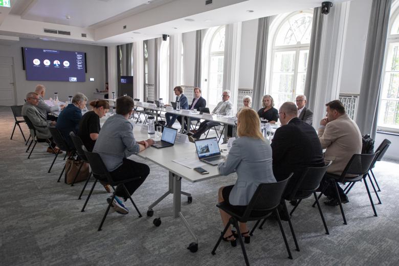 Członkowie Komisji rozmawiają siedząc przy stole konferencyjnym. W szczycie stołu znajduje się monitor wyświetlający członków Komisji łączących się ze spotkaniem za pośrednictwem Internetu