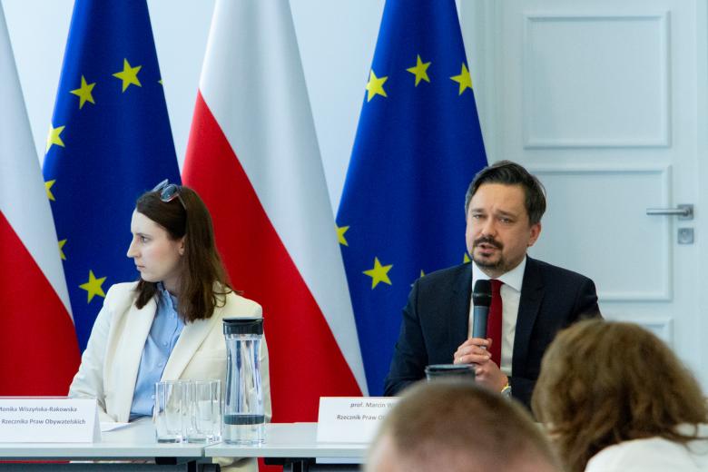 RPO Marcin Wiącek i Monika Wiszyńska-Rakowska siedzą przy stole konferencyjnym. Marcin Wiącek wypowiada się do mikrofonu w stronę uczestników spotkania siedzących przed nim, poza kadrem.