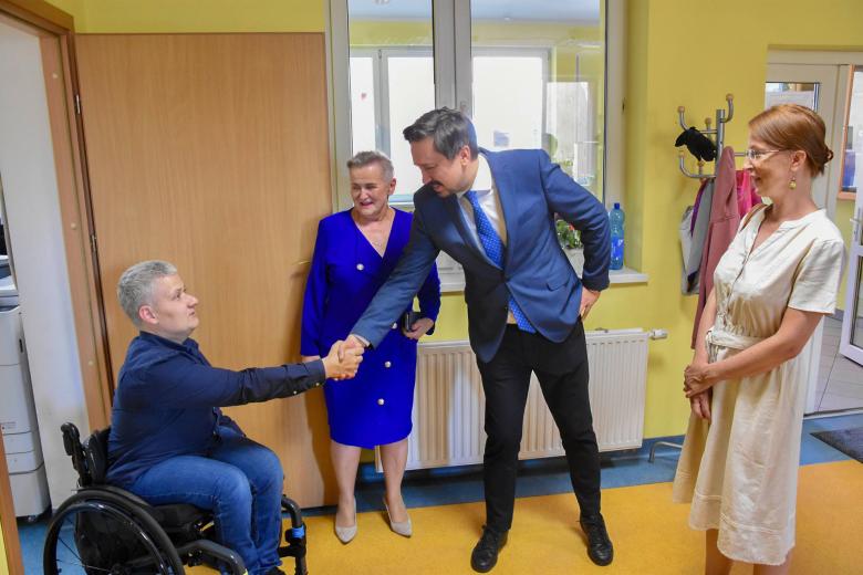 Mężczyzna wita się podaniem ręki z osobą poruszającą się na wózku inwalidzkim. Przyglądają się temu dwie kobiety.