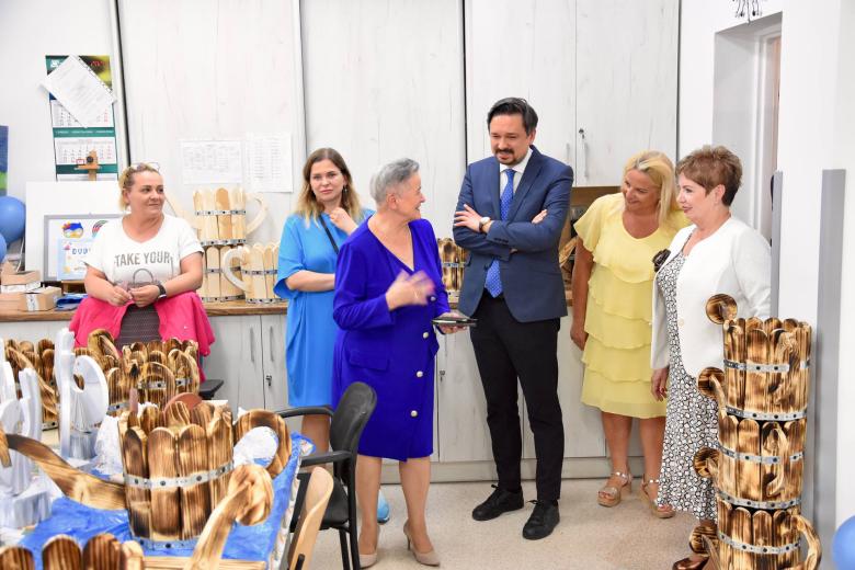 Sześć osób rozmawia w sali warsztatowej. Na stole stoją drewniane rękodzieła w kształcie konewek.