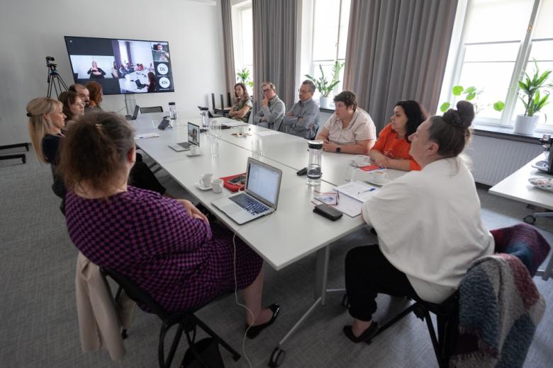 Członkowie Komisji rozmawiają przy konferencyjnym stole. W szczycie stołu znajduje się telewizor wyświetlający uczestników spotkania łączących się za pośrednictwem internetu oraz migającą tłumaczkę polskiego języka migowego.